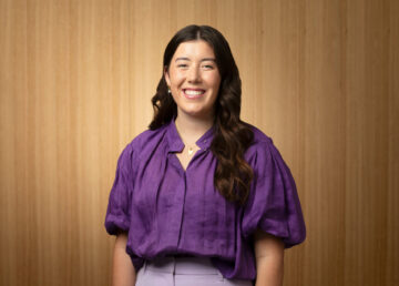 A woman with long, dark hair wearing a purple blouse and light purple pants smiles while standing in front of a wooden panel background.