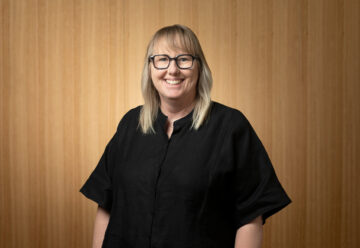 A person with shoulder-length blonde hair, glasses, and a black button-up shirt smiles while standing in front of a light wooden wall.