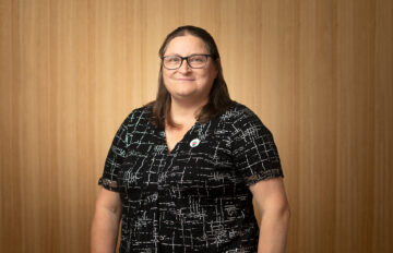 A woman with straight brown hair and glasses, wearing a black and white patterned blouse, stands in front of a wood-paneled background, smiling slightly at the camera.
