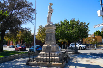 A statue of a soldier stands atop a stone war memorial inside a fenced area on a sidewalk, surrounded by trees and parked cars, with a clear blue sky overhead.