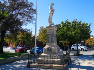 A stone war memorial statue of a soldier stands on a pedestal inscribed with names, surrounded by a black metal fence, trees, parked cars, and buildings on a sunny day. A street sign reads HARVEST RD.