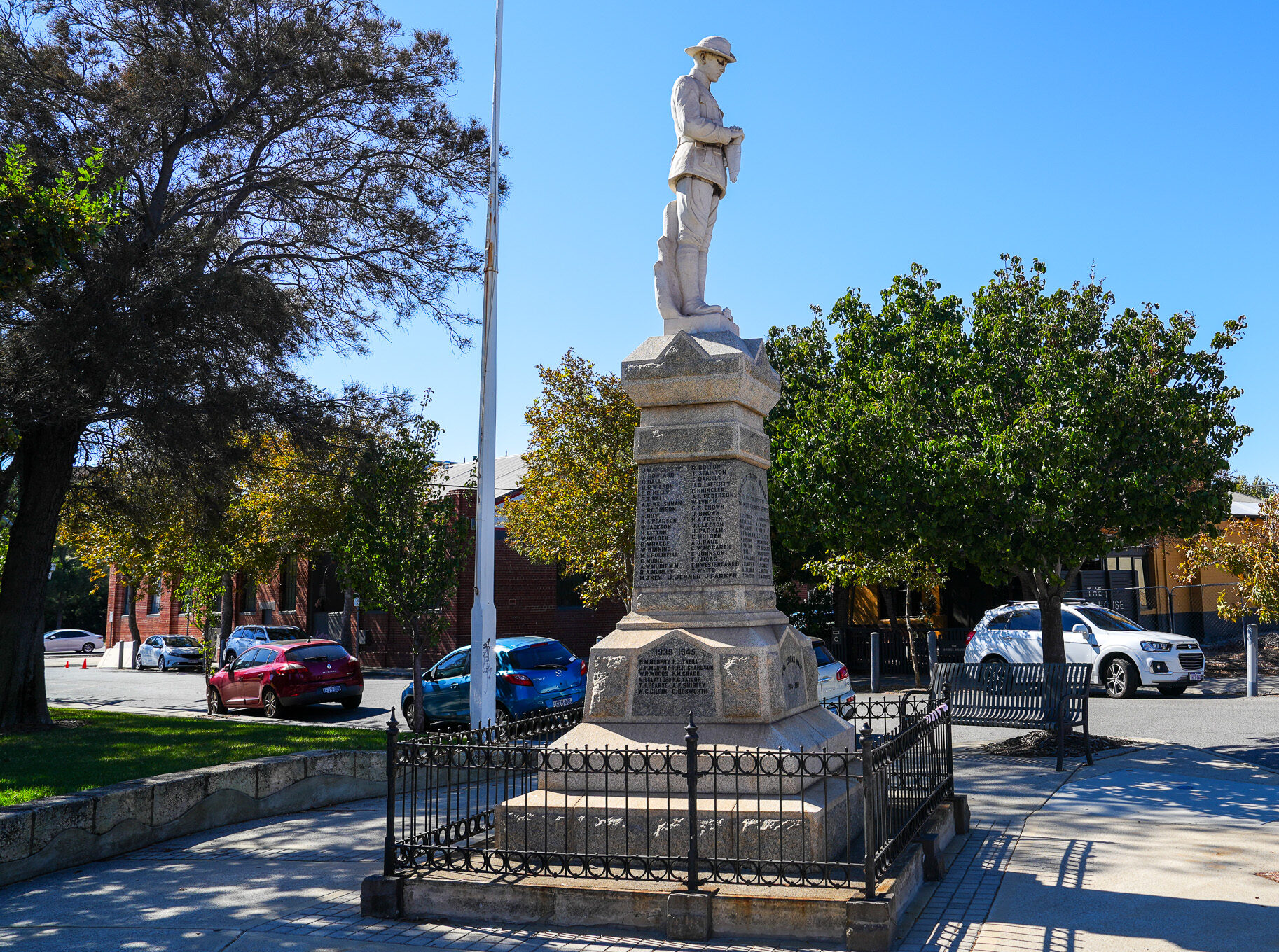 A stone war memorial statue of a soldier stands on a pedestal inscribed with names, surrounded by a black metal fence, trees, parked cars, and buildings on a sunny day. A street sign reads HARVEST RD.