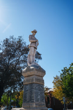 A stone war memorial statue of a soldier stands atop a tall pedestal inscribed with names, surrounded by trees and buildings under a clear blue sky.