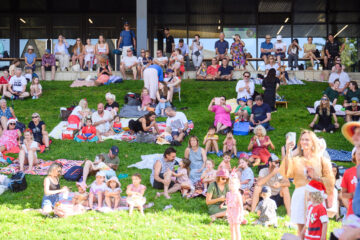A large group of people, including families with children, sit and relax on picnic blankets on a grassy hill in front of a modern building, enjoying a sunny outdoor event.