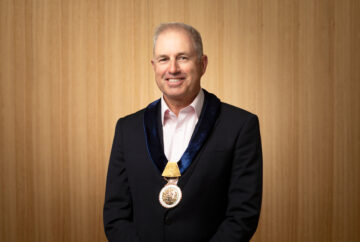 A man in a dark suit and light pink shirt stands in front of a wooden background, wearing a ceremonial gold medallion on a dark ribbon around his neck. He is smiling and facing the camera.