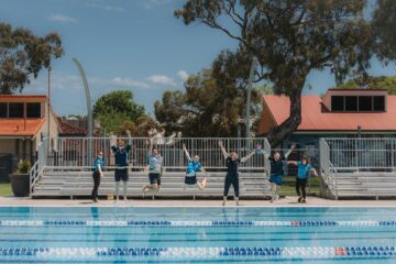 A group of people in sportswear jump joyfully in front of poolside bleachers at an outdoor swimming pool, with trees and buildings in the background on a sunny day.