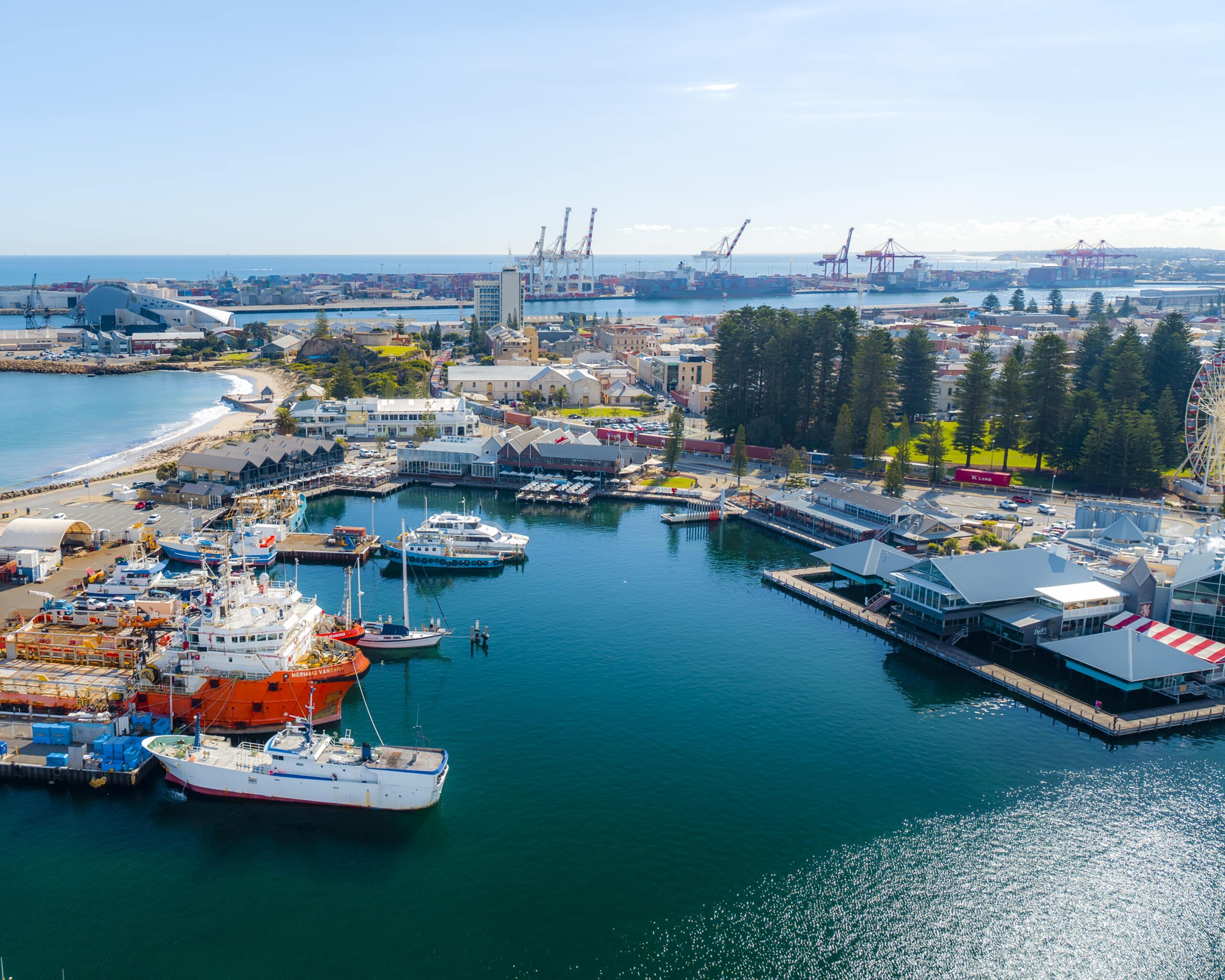 Aerial view of a harbor with several boats docked, waterfront buildings, a ferris wheel, and industrial cranes in the background under a clear blue sky.