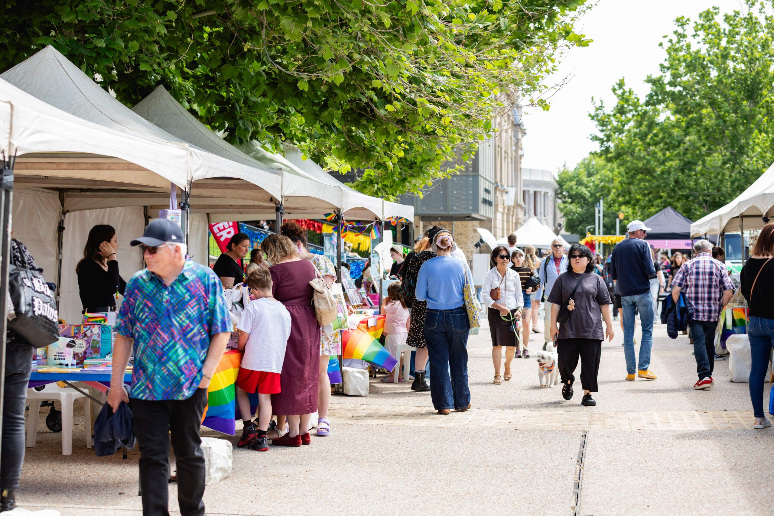 People walk along an outdoor market lined with white tents. Stalls display colorful goods, including rainbow flags. Some people chat or browse, while leafy trees provide shade along the path.