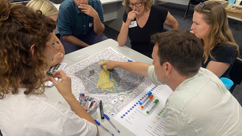 Six people sit around a table discussing and pointing at a large map covered with colored markers and notes, appearing to collaborate on urban planning or a community project.