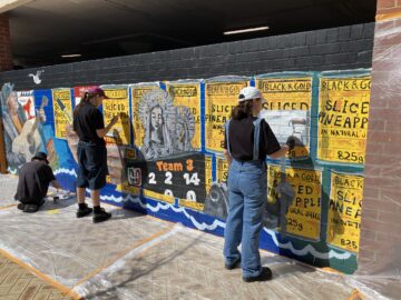 Three people paint a colorful mural on a brick wall featuring vintage-style pineapple can labels and an old ship. The ground is covered with plastic sheets to protect it from paint. Sunlight shines on the scene.