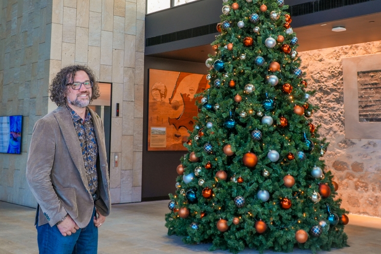 A man with curly hair, glasses, and a brown blazer stands smiling next to a large decorated Christmas tree in a modern indoor space with stone walls.