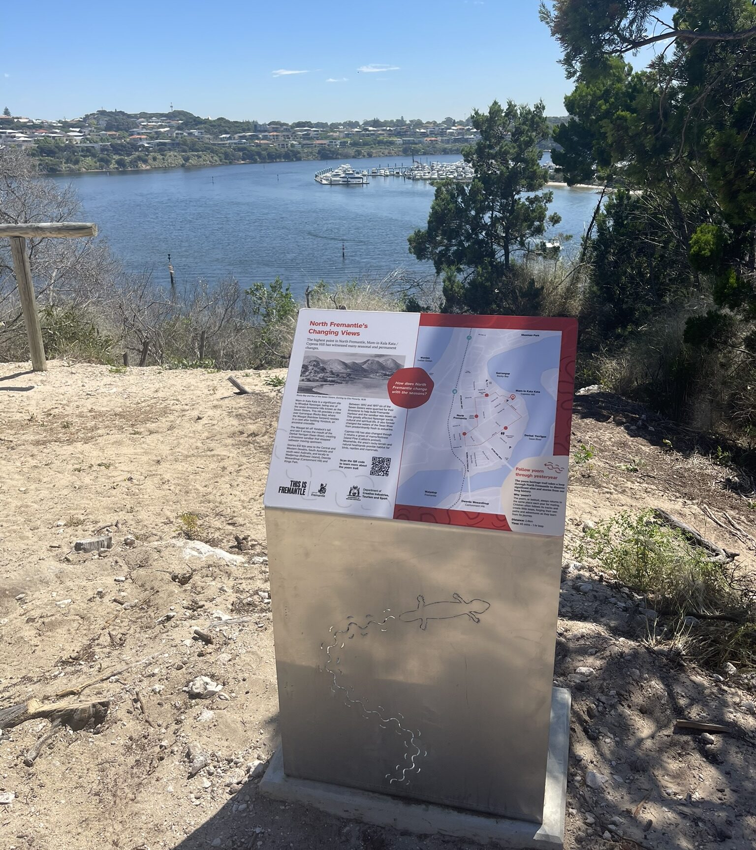 A metal information sign with a map and text stands on a sandy lookout, overlooking a river and suburban houses across the water, with trees partially framing the view under a clear blue sky.