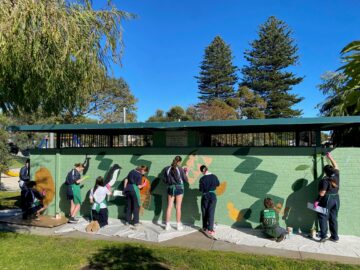 A group of children and teenagers paint a colorful mural with leaves on a green outdoor wall, standing on sheets to protect the grass. Trees and blue sky are visible in the background.