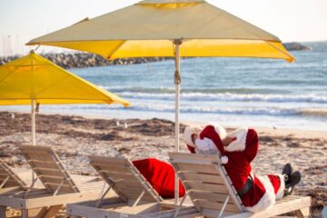 A person dressed as Santa Claus relaxes on a lounge chair under a yellow umbrella at a sandy beach, facing the ocean, with another empty lounge chair beside them.