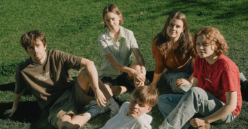 Five teenagers sit together on green grass in sunlight, looking up at the camera. They appear relaxed and friendly, dressed in casual summer clothes, with shadows cast on the ground around them.