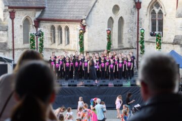 A choir of children and teens performs on an outdoor stage, wearing matching black shirts with pink designs. The audience, including children and adults, watches in front of a historic stone building decorated with Christmas garlands.