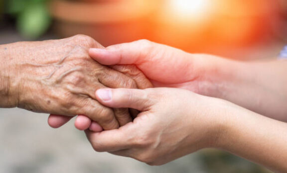 A younger person gently holding the hand of an older person, showing care and support; warm sunlight glows in the background.