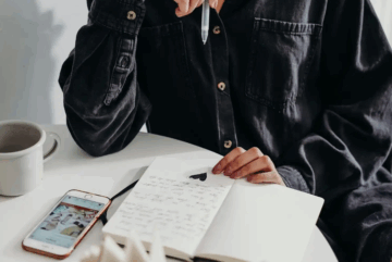 A person in a dark shirt sits at a table, holding a pen above an open notebook with handwritten text and a heart symbol. A cup and a smartphone displaying a website are nearby.