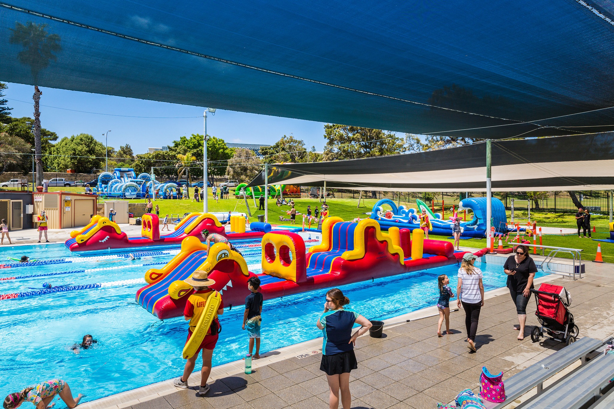 Children and adults enjoy an outdoor swimming pool with large, colorful inflatable obstacle courses. Some kids swim or climb the inflatables, while adults supervise from the poolside under shade cloths. Trees and grass are in the background.