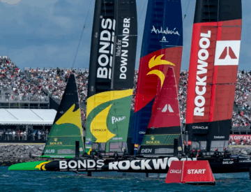 Several colorful racing sailboats with branding on their sails compete near a crowded spectator area by the water. A red buoy marked START floats in the foreground, and the sky is partly cloudy.