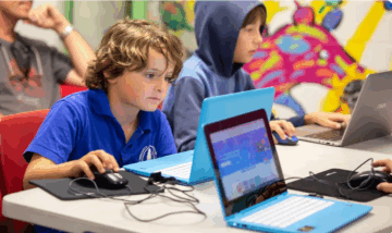 Two boys sit at a table using laptops and mice, focused on their screens. One wears a blue shirt, the other a hoodie. A colorful mural is visible in the background.