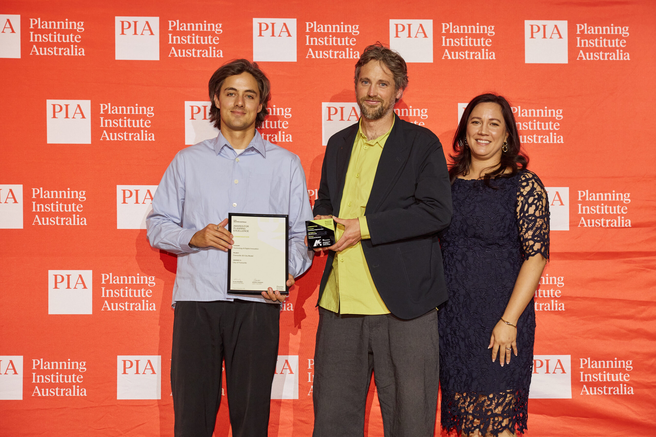 Three people stand in front of a red backdrop with Planning Institute Australia and PIA logos. Two men hold awards and a woman in a navy dress stands beside them, all smiling at the camera.