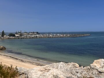A rocky shoreline overlooks a calm bay with clear blue water. A marina with numerous boats and yachts is visible, surrounded by a stone breakwater. Buildings and trees line the shore under a clear blue sky.