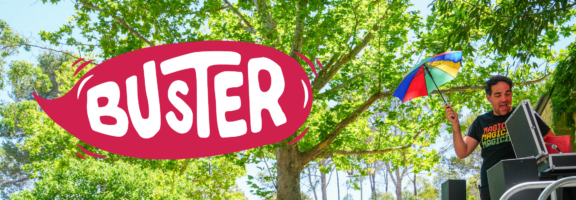A person holding a small rainbow umbrella stands beside equipment outdoors under green trees. Large, bold text BUSTER in white appears on a red shape to the left of the image.