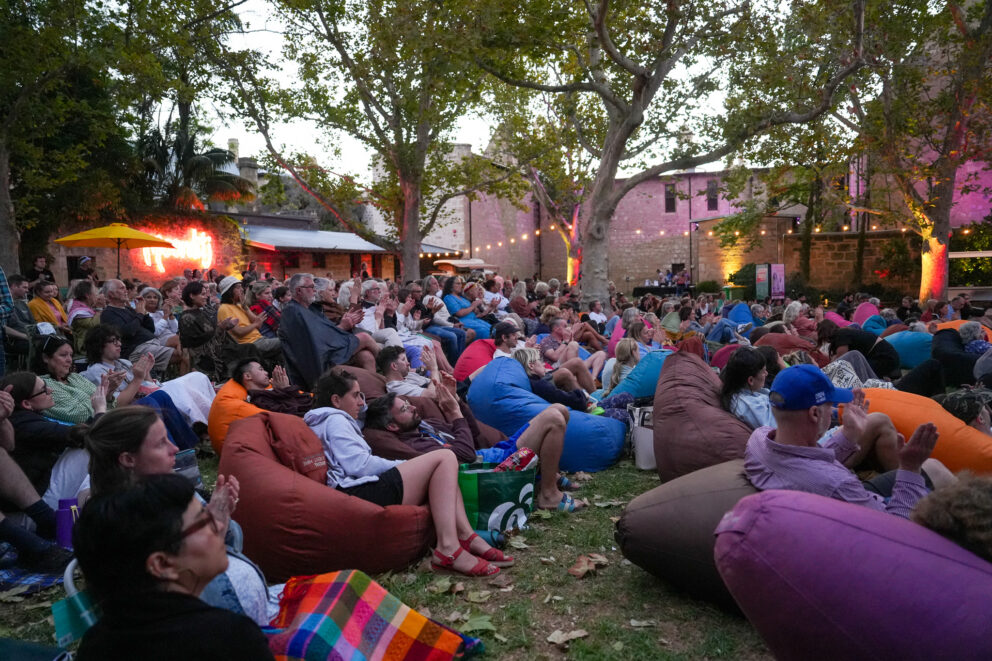 A large crowd of people sits on colorful bean bags and blankets outdoors, watching an event in a park-like setting with trees, string lights, and buildings in the background at dusk.