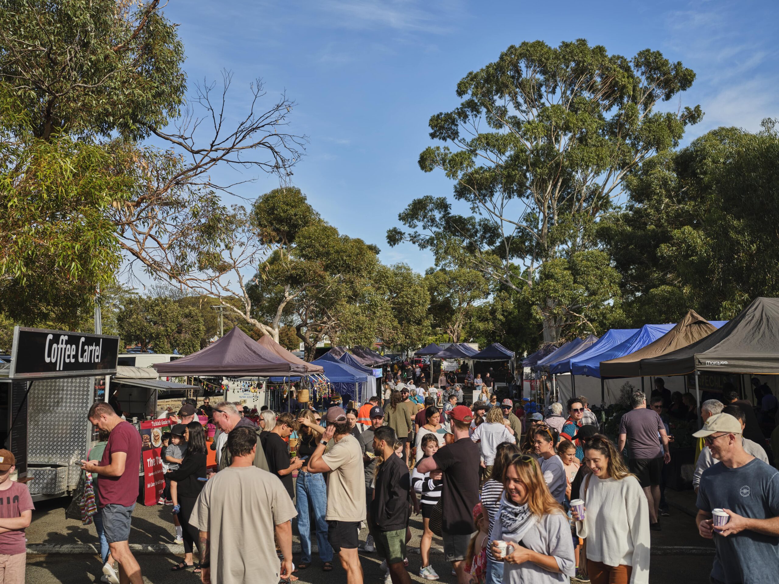 A busy outdoor market scene with many people walking among stalls and tents under tall trees. The crowd includes adults and children enjoying a sunny day. A food truck labeled “Coffee Cartel” is visible on the left.