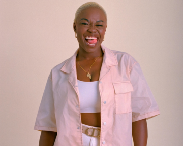A smiling woman with short blond hair wears a light pink shirt over a white crop top and pants. She stands against a neutral background, eyes closed, and sticks out her tongue playfully.