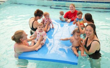 Four women sit in a pool with babies, playing together on a large blue float. Two women are on either side of the float, each holding a baby, while another woman and a child sit on the float. Bright, cheerful atmosphere.