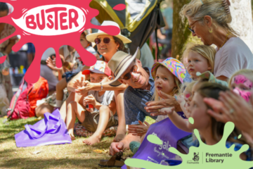 A group of adults and children sit on grass outdoors, smiling and clapping. Colorful banners with the words “Buster” and “Fremantle Library” are overlaid on the image. Sunlight filters through the trees.
