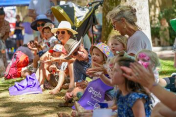A group of adults and children sitting on the grass at an outdoor event, some clapping and wearing hats.