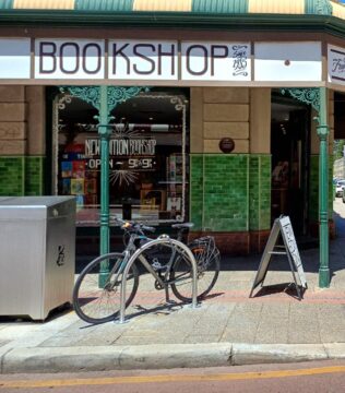 A sunny street corner with a vintage bookshop, a bicycle parked outside, and pedestrians walking nearby. The bookshop has green tiles, large windows, and signs for new editions and rare books. Historic buildings line the street.