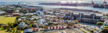 Aerial view of a coastal city with residential houses, apartment buildings, green spaces, and a busy port with cargo ships docked along the waterfront under a bright, sunny sky.