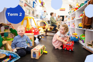 Two young children play with toys on the floor of a colorful, well-stocked toy store or playroom. Adults and other children are in the background. A blue circle in the corner reads Term 2.