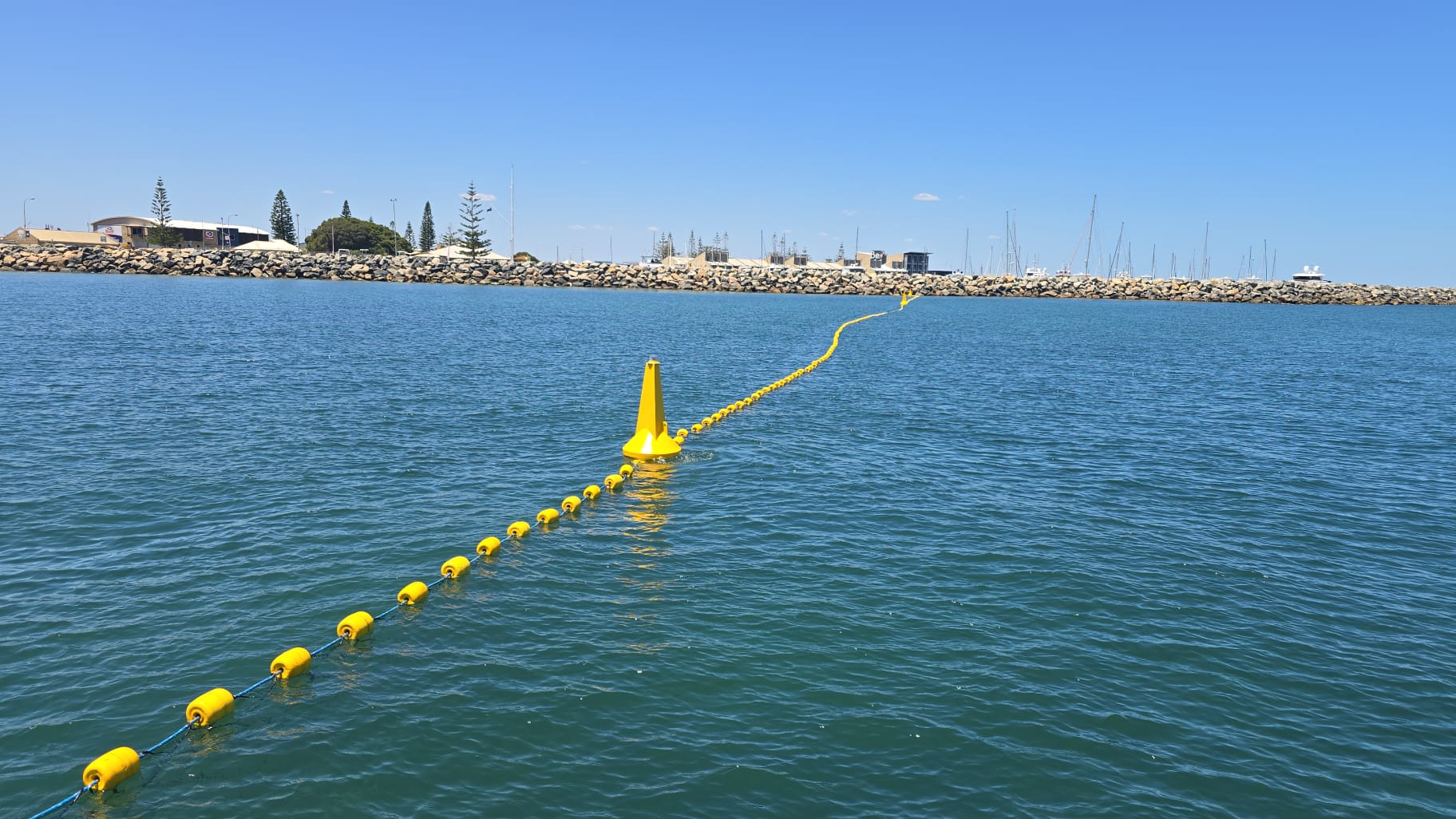 A line of yellow buoys floats on calm blue water, leading to a yellow cone marker. In the background, a rocky breakwater and a marina with sailboats and buildings are visible under a clear blue sky.