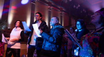 A group of people, holding sheets of paper, sing together in a dimly lit room with colorful, dramatic lighting and high ceilings.