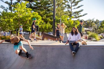 Children skateboard at a sunny park with trees in the background, while a woman sits on the edge of the skate bowl, smiling and holding a clipboard. Other kids watch and play nearby.