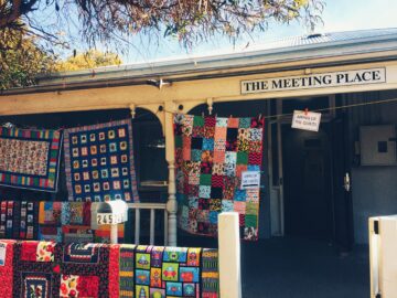 Colorful patchwork quilts hang on the porch and fence of a building called The Meeting Place, with a sign reading Airing of the Quilts under a sunny, tree-shaded sky.