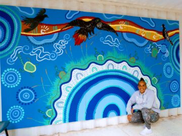 A person in a grey sweatshirt kneels and smiles in front of a vibrant blue mural featuring circular Aboriginal patterns, turtles, and three black birds flying near a red-and-yellow wavy line.