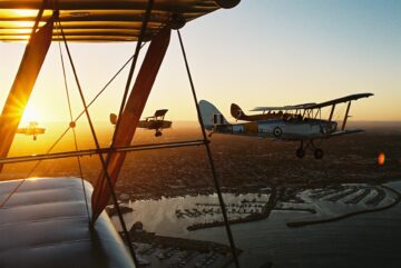 Four vintage biplanes fly in formation over a coastal city at sunset, with the sun casting a warm glow over the scene and a harbor visible below.