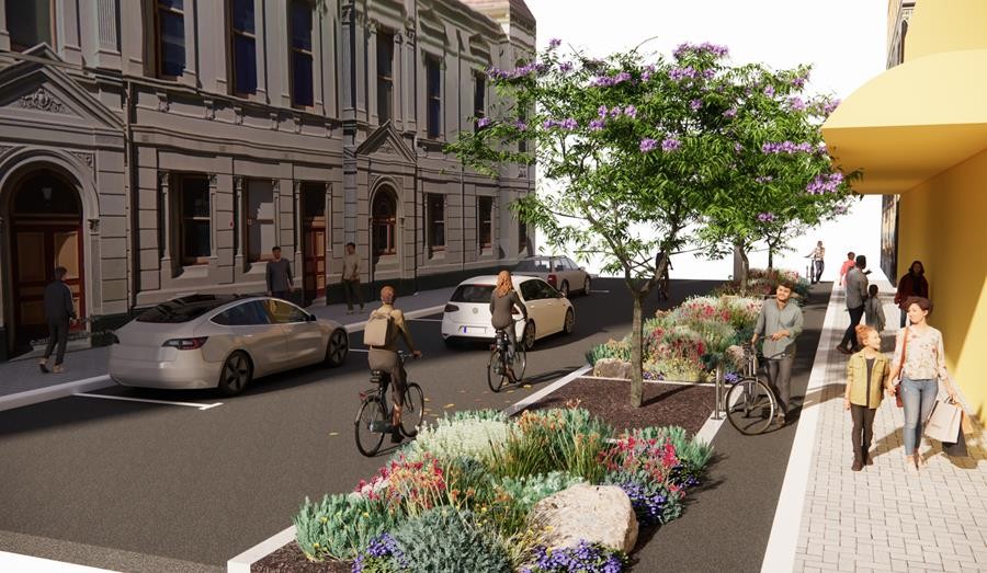 A city street with cyclists on a bike lane, pedestrians walking on a wide sidewalk, and cars parked along the curb. Trees and colorful plants line the sidewalk, with historic buildings in the background.