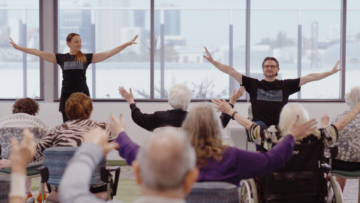 Two instructors lead a group of seniors in seated exercise, all raising their arms. The class takes place indoors, with large windows overlooking a city skyline in the background.