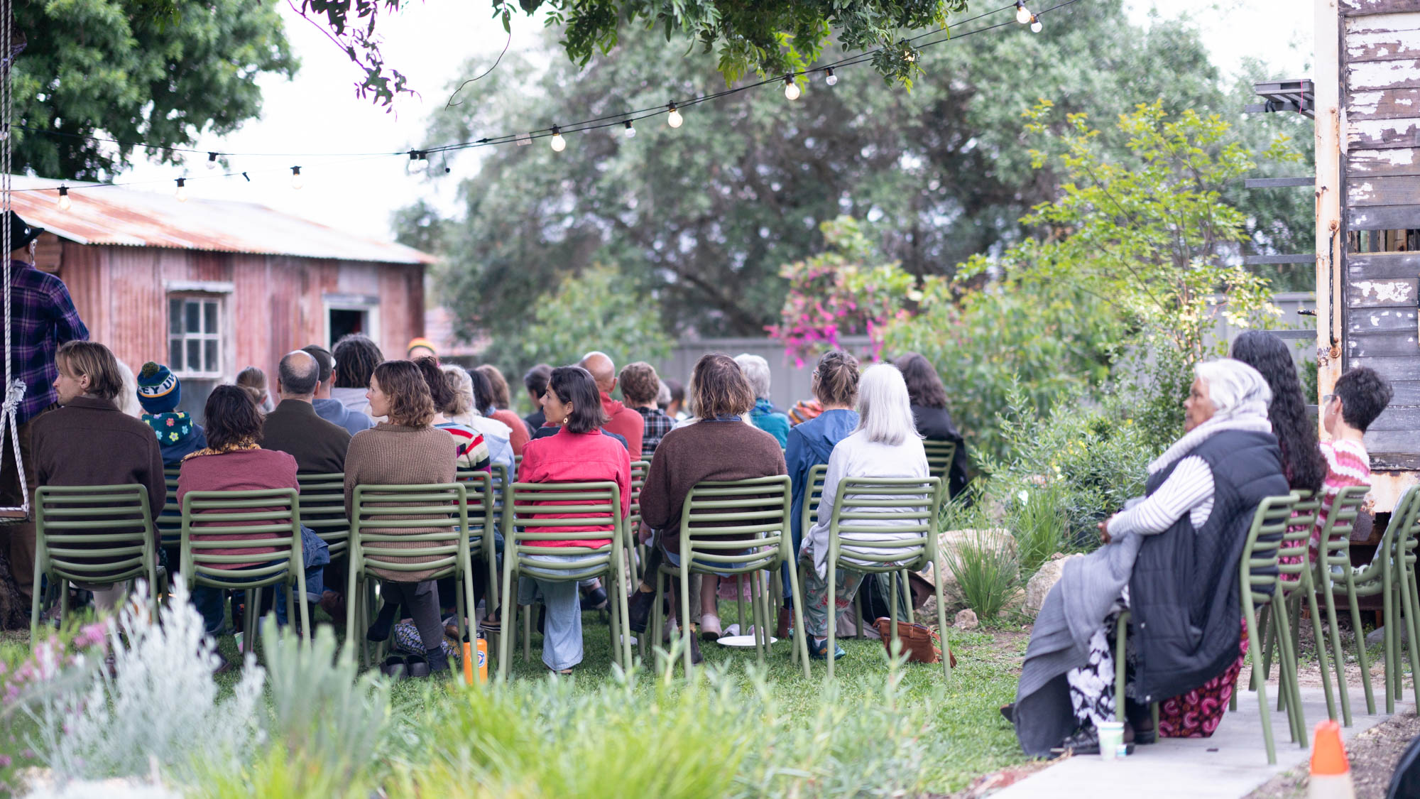 A group of people sitting on green chairs outdoors, listening to a speaker at an event. String lights hang above, and there is greenery and rustic buildings in the background.