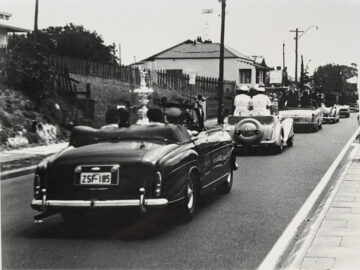 A black-and-white photo of a vintage car parade driving on a suburban street; the lead convertible carries a large trophy, followed by several classic cars and people in formal attire.