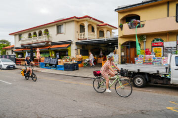Two people ride bicycles past a local market with fresh produce and goods on display outside. A white truck is parked in front of colorful store buildings on a cloudy day.