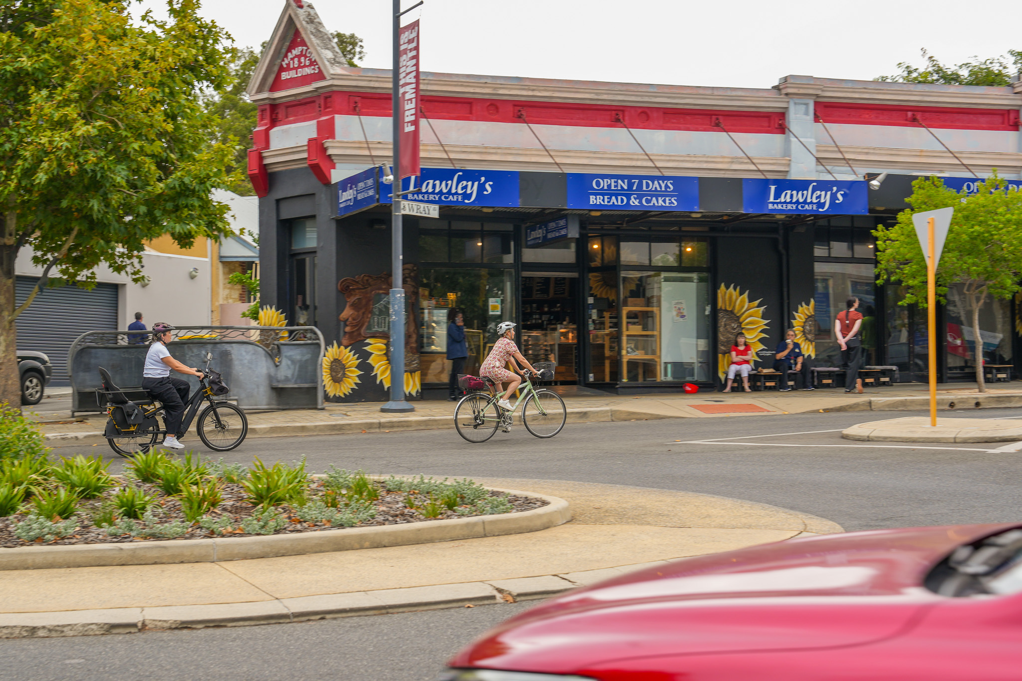 Two people ride bicycles past a bakery with sunflower murals on the wall. A red car passes in the foreground, and a few people sit outside the bakery. Trees and a small garden are visible around the street corner.