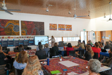 A group of people sit at tables covered with colorful cloths, listening to a presenter speaking in front of a screen in a brightly lit room decorated with Indigenous Australian art.